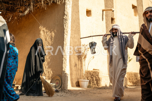مهنة شعبية متوارثة، ذكرى تأسيس الدولة السعودية الأولى1727 م، بيت طيني مغطى بسعائف الخوص الجاف، قرية أثرية تاريخية قديمة، مجموعة أشخاص يتجولون في سوق شعبي، يوم بدينا22 فبراير، رجل عربي خليجي سعودي يرتدي الثوب التقليدي والغترة يحمل دلوين بواسطة عصا خشبية طويلة