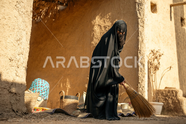 يوم بدينا22 فبراير، ذكرى تأسيس الدولة السعودية الأولى1727 م،  امرأة عربية خليجية سعودية ترتدي العباءة والنقاب تكنس بمكنسة من القش في سوق شعبي قديم, المحافظة على التراث السعودي