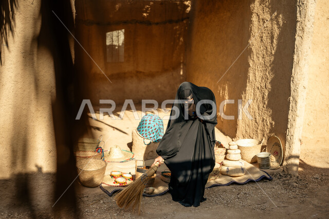 يوم بدينا22 فبراير، ذكرى تأسيس الدولة السعودية الأولى1727 م،  امرأة عربية خليجية سعودية ترتدي العباءة والنقاب تكنس بمكنسة من القش في سوق شعبي قديم, المحافظة على التراث السعودي