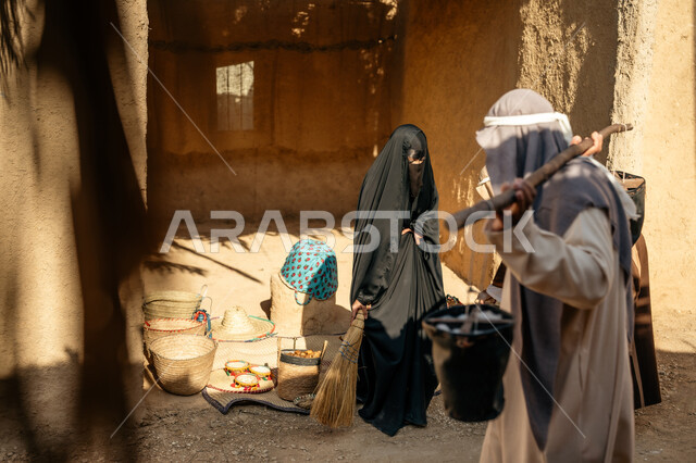 ذكرى تأسيس الدولة السعودية الأولى1727 م، قرية أثرية تاريخية قديمة، مهنة شعبية متوارثة، يوم بدينا22 فبراير، صورة من الجانب لرجل عربي خليجي سعودي يرتدي الثوب التقليدي والغترة يحمل دلوين بواسطة عصا خشبية طويلة, امرأة عربية خليجية سعودية ترتدي العباءة والنقاب تكنس بمكنسة من القش