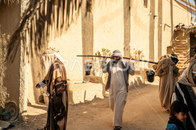 مهنة شعبية متوارثة، ذكرى تأسيس الدولة السعودية الأولى1727 م، بيت طيني مغطى بسعائف الخوص الجاف، قرية أثرية تاريخية قديمة، مجموعة أشخاص يتجولون في سوق شعبي، يوم بدينا22 فبراير، رجل عربي خليجي سعودي يرتدي الثوب التقليدي والغترة يحمل دلوين بواسطة عصا خشبية طويلة