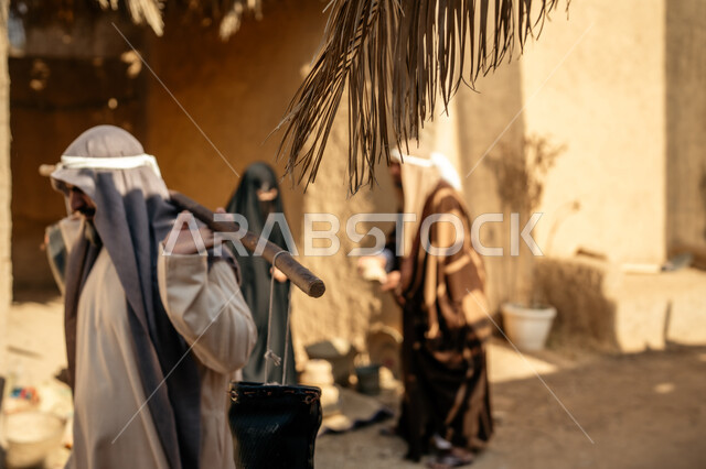 قرية أثرية تاريخية قديمة، مهنة شعبية متوارثة، ذكرى تأسيس الدولة السعودية الأولى1727 م، بيت طيني مغطى بسعائف الخوص الجاف، مجموعة أشخاص يتجولون في سوق شعبي، يوم بدينا22 فبراير، رجل عربي خليجي سعودي يرتدي الثوب التقليدي والغترة يحمل دلوين بواسطة عصا خشبية طويلة