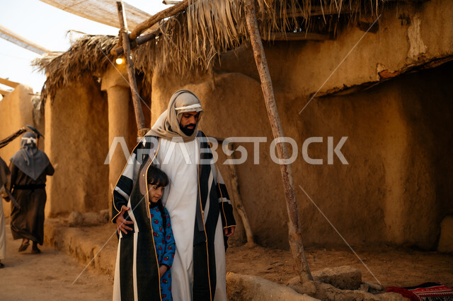 Providing shade in an old, inherited way, a cloth canopy extended between the houses, on the Day of Our Day, February 22, the anniversary of the founding of the first Saudi state, 1727 AD. A Saudi Gulf Arab man wearing a bisht and ghutra covers his daughter and they walk around the historic neighborhood built in the old style.