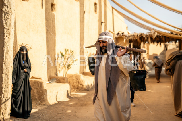 قرية أثرية تاريخية قديمة، مهنة شعبية متوارثة، ذكرى تأسيس الدولة السعودية الأولى1727 م، بيت طيني مغطى بسعائف الخوص الجاف، مجموعة أشخاص يتجولون في سوق شعبي، يوم بدينا22 فبراير، رجل عربي خليجي سعودي يرتدي الثوب التقليدي والغترة يحمل دلوين بواسطة عصا خشبية طويلة