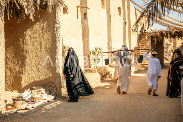 يوم بدينا22 فبراير، مهنة شعبية متوارثة، ذكرى تأسيس الدولة السعودية الأولى1727 م، بيت طيني مغطى بسعائف الخوص الجاف، صبي يمشي في سوق شعبي يقوم بتقديم المساعدة، صورة من الخلف لرجل عربي خليجي سعودي يرتدي الثوب التقليدي والغترة يحمل دلوين بواسطة عصا خشبية طويلة