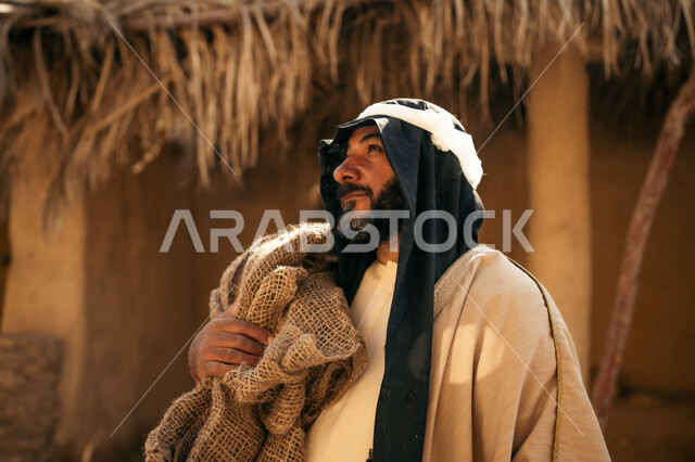 ذكرى تأسيس الدولة السعودية الأولى1727م, خلفية سقف المنزل الطيني المغطى بسعف النخيل الجاف, يوم بدينا22 فبراير, قرية تاريخية تراثية، صورة مقربة لرجل عربي خليجي سعودي يرتدي البشت والغترة يقف أمام بيت طيني قديم يحمل كيس قماشي وينظر لمكان ما بايماءات الفخر