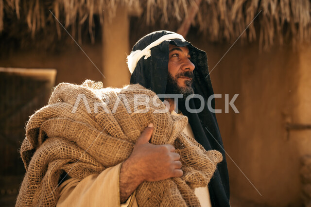 ذكرى تأسيس الدولة السعودية الأولى1727م, خلفية بيت من الطين مغطى بسعف النخل, يوم بدينا22 فبراير, قرية تاريخية تراثية، صورة مقربة لرجل عربي خليجي سعودي يرتدي البشت والغترة  يحمل كيس قماشي وينظر لمكان ما بايماءات الفخر