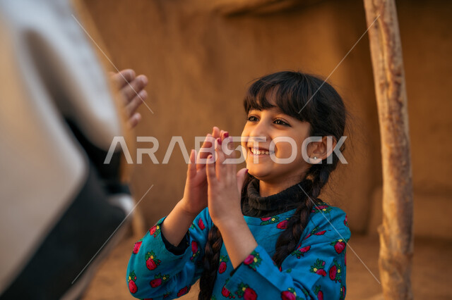 ألعاب متوارثة من الأجداد عبر الأجيال، قضاء وقت ممتع مع الأب، بنت عربية خيليجية سعودية ترتدي فستان أزرق تقوم باللعب والمرح برفقة والدها بإيماءات السعادة والسرور، إحياء ذكرى تأسيس الدولة السعودية الأولى 1727 م, يوم بدينا 22 فبراير, منزل طيني قديم في قرية تاريخية