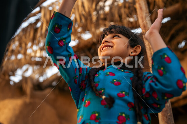 ألعاب متوارثة من الأجداد عبر الأجيال، قضاء وقت ممتع مع الأب، بنت عربية خيليجية سعودية ترتدي فستان أزرق تقوم باللعب والمرح برفقة والدها بإيماءات السعادة والسرور، إحياء ذكرى تأسيس الدولة السعودية الأولى 1727 م, يوم بدينا 22 فبراير, منزل طيني قديم في قرية تاريخية