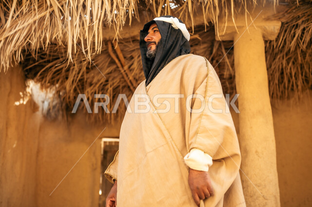 ارتداء الزي الشعبي في مناسبة وطنية, قرية تاريخية تراثية، يوم بدينا22 فبراير, ذكرى تأسيس الدولة السعودية الأولى1727م, رجل عربي خليجي سعودي يرتدي الثوب التقليدي ويضع البشت والغترة يقف أمام بيت طيني قديم بايماءات الفخر، خلفية سقف المنزل المغطى بسعف النخل