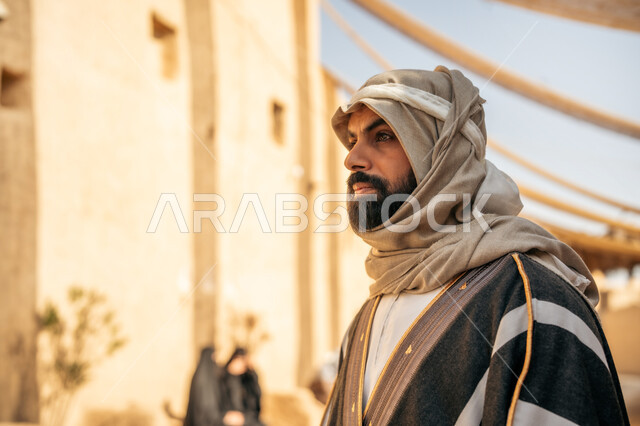 ذكرى تأسيس الدولة السعودية الأولى1727م, يوم بدينا22 فبراير, قرية تاريخية تراثية، ارتداء الزي الشعبي في مناسبة وطنية, رجل عربي خليجي سعودي يرتدي الثوب التقليدي ويضع البشت والغترة يقف أمام بيت طيني قديم بايماءات الفخر
