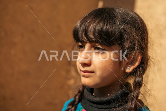 مفهوم النعومة والبراءة, إحياء ذكرى يوم التأسيس السعودي 1727 م, يوم بدينا 22 فبراير, صورة مقربة لفتاة عربية خليجية سعودية ترتدي فستان أزرق تقف أمام بيت من الطين  وتنظر إلى مكان ما بإيماءات السعادة و السرور, الأناقة والاهتمام بالمظهر الخارجي