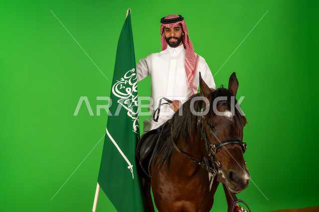 الفخر والاعتزاز بالهوية الوطنية، شغف تربية ورعاية الخيل، توارث حب الجياد من الاجداد، اليوم الوطني السعودي 23 سبتمبر، يوم العلم 11 مارس، بورتريه لرجل عربي خليجي سعودي يرتدي الثوب التقليدي والشماغ يمتطي فرس عربي يرفع علم السعودية، خلفية خضراء، كروما