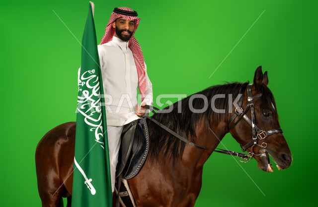 الفخر والاعتزاز بالهوية الوطنية، شغف تربية ورعاية الخيل، توارث حب الجياد من الاجداد، اليوم الوطني السعودي 23 سبتمبر، يوم العلم 11 مارس، بورتريه لرجل عربي خليجي سعودي يرتدي الثوب التقليدي والشماغ يمتطي فرس عربي يرفع علم السعودية، خلفية خضراء، كروما