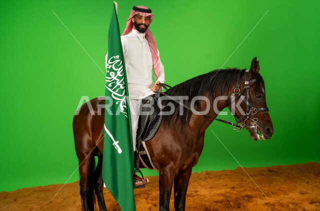 الفخر والاعتزاز بالهوية الوطنية، يوم العلم 11 مارس، بورتريه لرجل عربي خليجي سعودي يرتدي الثوب التقليدي والشماغ يمتطي فرس عربي يرفع علم السعودية فوق الرمال الناعمة، توارث حب الجياد من الاجداد، اليوم الوطني السعودي 23 سبتمبر، شغف تربية ورعاية الخيل، خلفية خضراء، كروما