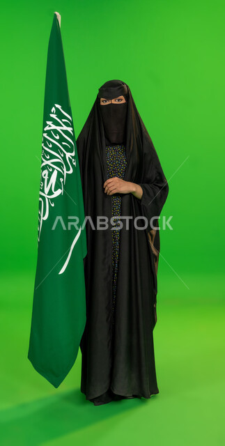يوم بدينا 22 فبراير, ذكرى تأسيس الدولة السعودية الأولى1727م, اليوم الوطني السعودي 23 سبتمبر، بورتريه لامرأة عربية خليجية سعودية ترتدي البرقع والعباءة تمسك علم المملكة تنظر إلى الكاميرا، يوم العلم 11 مارس، حب الوطن والانتماء إليه، الجسم بالطول الكامل، خلفية خضراء، كروما