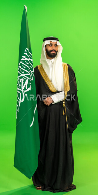 ارتداء الزي الشعبي في مناسبة وطنية, ذكرى تأسيس الدولة السعودية الأولى1727م, اليوم الوطني السعودي 23 سبتمبر، يوم العلم 11 مارس، يوم بدينا22 فبراير, بورتريه لرجل عربي خليجي سعودي يرتدي البشت والغترة يمسك علم المملكة، الطول الكامل للجسم، خلفية خضراء، كروما