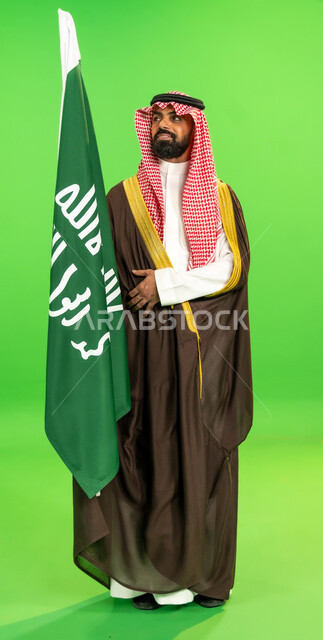 الفخر والاعتزاز بالانتماء للوطن, يوم بدينا22 فبراير, ذكرى تأسيس الدولة السعودية الأولى1727م, ارتداء الزي الشعبي في مناسبة وطنية, بورتريه لرجل عربي خليجي سعودي يرتدي البشت والشماغ يمسك علم يوم التأسيس بتعابير الشموخ ، الطول الكامل للجسم، خلفية خضراء، كروما