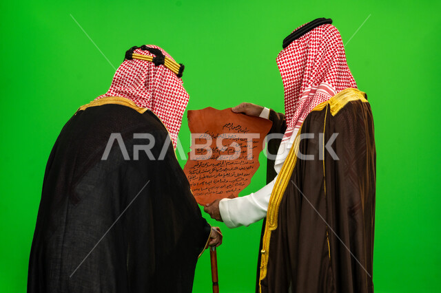 أحياء تراث المملكة, يوم بدينا 22 فبراير, ذكرى تأسيس الدولة السعودية الأولى 1727م، العراقة والاصالة عند العرب، ارتداء الزي التقليدي في مناسبة وطنية، بورتريه مقرب من الخلف لرجلين عربيين خليجيين سعوديين يرتديان البشت والشماغ يقومان بقراءة مخطوطة جلد تاريخية، خلفية خضراء، كروما
