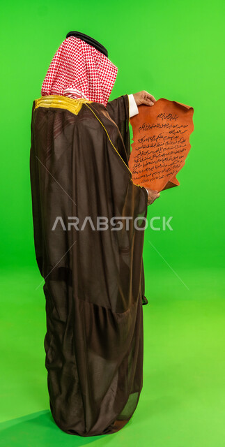العراقة والاصالة عند العرب، ذكرى تأسيس الدولة السعودية الأولى 1727م، ارتداء الزي التقليدي في مناسبة وطنية, يوم بدينا 22 فبراير, بورتريه من الخلف لرجل عربي خليجي سعودي يرتدي البشت والشماغ يقوم بقراءة مخطوطة جلد تاريخية، الطول الكامل للجسم، خلفية خضراء، كروما