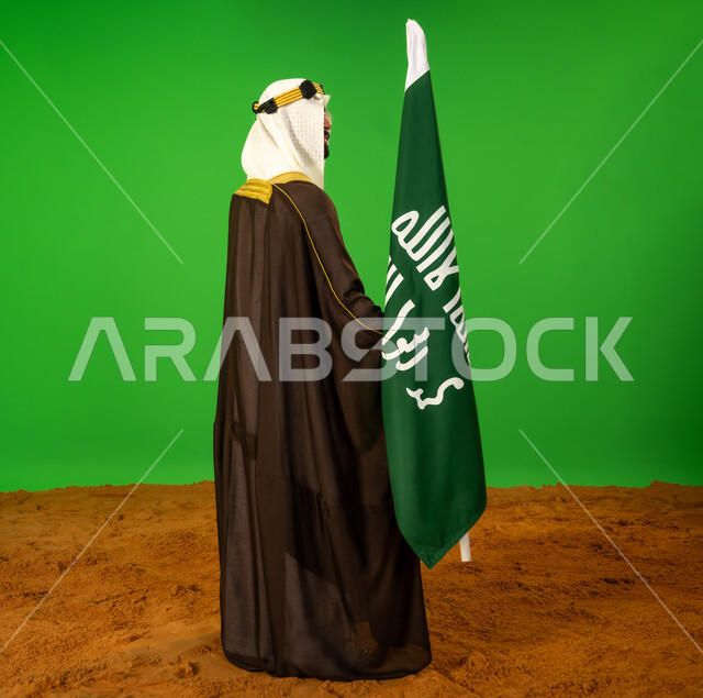 يوم بدينا22 فبراير، ذكرى تأسيس الدولة السعودية الأولى1727م, حب الوطن والانتماء اليه، ارتداء الزي الشعبي في مناسبة وطنية, بورتريه من الجانب لرجل عربي خليجي سعودي يرتدي البشت والعقال يمسك علم يوم التأسيس ويقف على الرمال الناعمة، الطول الكامل للجسم، خلفية خضراء، كروما