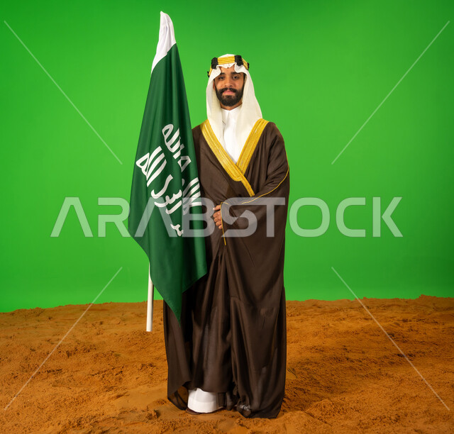 الاعتزاز والفخر بالهوية الوطنية، يوم بدينا22 فبراير, ذكرى تأسيس الدولة السعودية الأولى1727م, ارتداء الزي الشعبي في مناسبة وطنية, بورتريه لرجل عربي خليجي سعودي يرتدي البشت والعقال يمسك علم يوم التأسيس ويقف على الرمال الناعمة، الطول الكامل للجسم، خلفية خضراء، كروما
