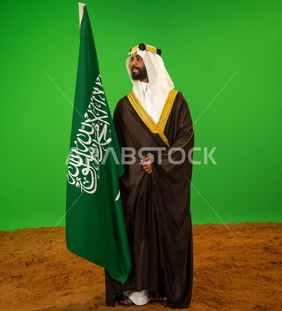 ذكرى تأسيس الدولة السعودية الأولى1727م, يوم بدينا22 فبراير, الاعتزاز والفخر بالهوية الوطنية، ارتداء الزي الشعبي في مناسبة وطنية, بورتريه لرجل عربي خليجي سعودي يرتدي البشت والعقال يمسك علم يوم التأسيس ويقف على الرمال الناعمة، الطول الكامل للجسم، خلفية خضراء، كروما