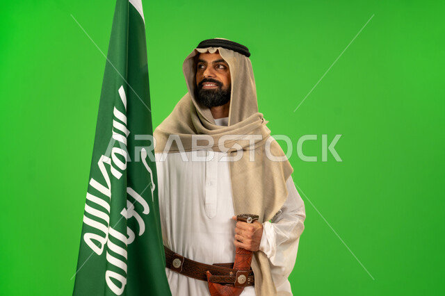 ذكرى تأسيس الدولة السعودية الأولى1727م, يوم بدينا22 فبراير, مفهوم حب الوطن والانتماء اليه, ارتداء الزي الشعبي في مناسبة وطنية, بورتريه لرجل عربي خليجي سعودي يرتدي الثوب التقليدي والعقال يضع الحزام والخنجر يمسك علم يوم التأسيس وينظر اليه بتعابير شموخ، خلفية خضراء، كروما