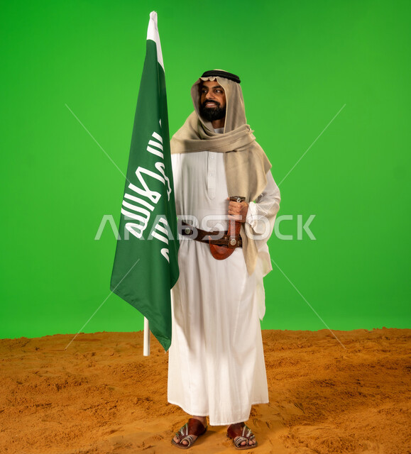 ارتداء الزي الشعبي في مناسبة وطنية, ذكرى تأسيس الدولة السعودية الأولى1727م, يوم بدينا22 فبراير, حب الوطن والانتماء اليه, بورتريه لرجل عربي خليجي سعودي يرتدي الثوب التقليدي والعقال يضع الحزام والخنجر يقف على الرمال يمسك علم يوم التأسيس، الطول الكامل للجسم، خلفية خضراء، كروما