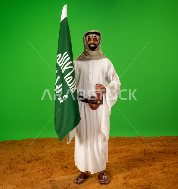 ذكرى تأسيس الدولة السعودية الأولى1727م, يوم بدينا22 فبراير, حب الوطن والانتماء اليه, ارتداء الزي الشعبي في مناسبة وطنية, بورتريه لرجل عربي خليجي سعودي يرتدي الثوب التقليدي والعقال يضع الحزام والخنجر يقف على الرمال يمسك علم يوم التأسيس، الطول الكامل للجسم، خلفية خضراء، كروما