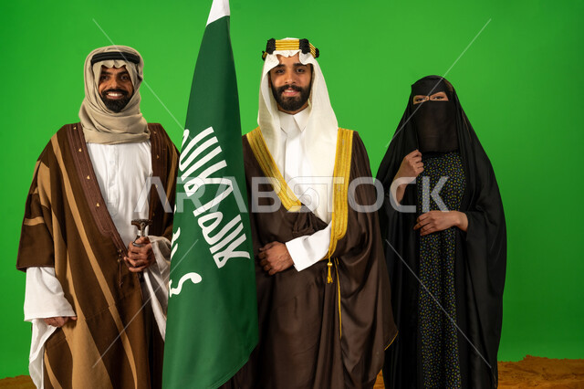 النظر للكاميرا بايماءات الفخر، يوم بدينا 22 فبراير، بورتريه لأشخاص عرب خليجيين سعوديين يرتدون الزي الشعبي التقليدي في مناسبة وطنية يقفون على الرمال الناعمة، رجل سعودي يحمل علم يوم التأسيس، الاصالة والعراقة عند العرب، ذكرى تأسيس الدولة السعودية الأولى 1727م، خلفية خضراء، كروما