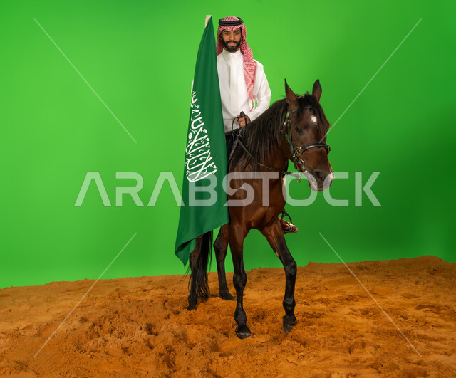 يوم العلم 11 مارس، الفخر والاعتزاز بالهوية الوطنية، شغف تربية ورعاية الخيل، توارث حب الجياد من الاجداد، اليوم الوطني السعودي 23 سبتمبر، بورتريه لرجل عربي خليجي سعودي يرتدي الثوب التقليدي والشماغ يمتطي فرس عربي ويرفع علم السعودية فوق الرمال الناعمة، خلفية خضراء، كروما