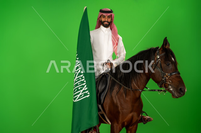 الفخر والاعتزاز بالهوية الوطنية، شغف تربية ورعاية الخيل، توارث حب الجياد من الاجداد، اليوم الوطني السعودي 23 سبتمبر، يوم العلم 11 مارس، بورتريه لرجل عربي خليجي سعودي يرتدي الثوب التقليدي والشماغ يمتطي فرس عربي ويرفع علم السعودية، خلفية خضراء، كروما