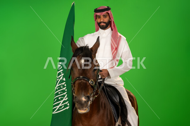 اليوم الوطني السعودي 23 سبتمبر، يوم العلم 11 مارس، الاعتزاز والفخر بالهوية الوطنية، شغف تربية ورعاية الخيل، توارث حب الجياد من الاجداد، بورتريه لرجل عربي خليجي سعودي يرتدي الثوب التقليدي والشماغ يمتطي فرس عربي ويرفع علم السعودية، خلفية خضراء، كروما