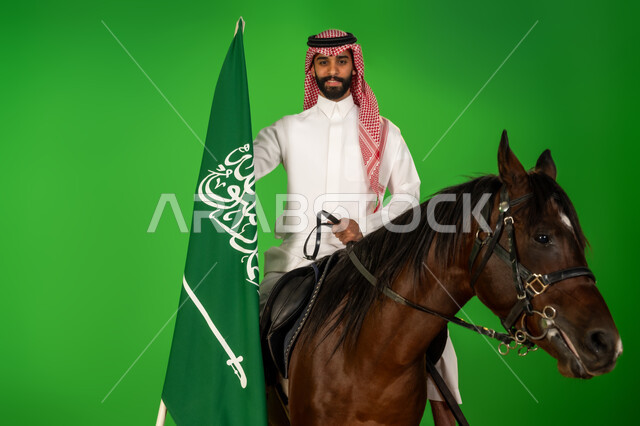 الاعتزاز والفخر بالهوية الوطنية، شغف تربية ورعاية الخيل، توارث حب الجياد من الاجداد، اليوم الوطني السعودي 23 سبتمبر، يوم العلم 11 مارس، بورتريه لرجل عربي خليجي سعودي يرتدي الثوب التقليدي والشماغ يمتطي فرس عربي ويرفع علم السعودية، خلفية خضراء، كروما