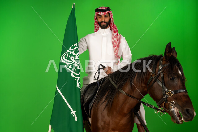 الفخر والاعتزاز بالهوية الوطنية، شغف تربية ورعاية الخيل، توارث حب الجياد من الاجداد، اليوم الوطني السعودي 23 سبتمبر، يوم العلم 11 مارس، بورتريه لرجل عربي خليجي سعودي يرتدي الثوب التقليدي والشماغ يمتطي فرس عربي ويرفع علم السعودية، خلفية خضراء، كروما