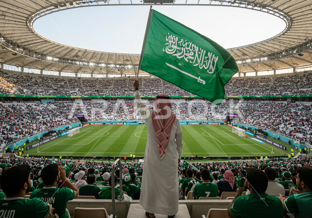 تشجيع منتخب السعودية، بطولة كأس العالم 2034، صورة من الخلف لرجل عربي خليجي سعودي يرتدي الثوب التقليدي والشماغ يحمل علم المملكة, الاهتمام بالرياضة والأنشطة الترفيهية، دعم الفرق والمنتخبات والأندية