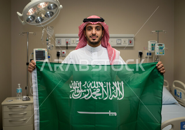 التقدم العمراني للمنشآت الطبية، يوم العلم 11 مارس، رجل عربي خليجي سعودي يرتدي الثوب التقليدي والشماغ يقف داخل غرفة في مشفى يحمل علم المملكة ينظر إلى الكاميرا بإيماءات الفخر، اليوم الوطني السعودي 23 سبتمبر, المراكز الصحية والمستشفيات وأماكن العناية بالمرضى