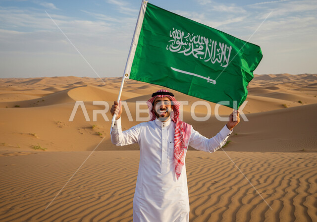 اليوم الوطني السعودي 23 سبتمبر, البادية والبيئة الصحراوية في النهار، يوم العلم 11 مارس، رجل عربي خليجي سعودي مبتسم يرتدي الثوب التقليدي والشماغ يقف على الرمال الناعمة في الصحراء يحمل علم المملكة ينظر إلى الكاميرا بإيماءات الفخر، التشكلات الرملية في الصحاري