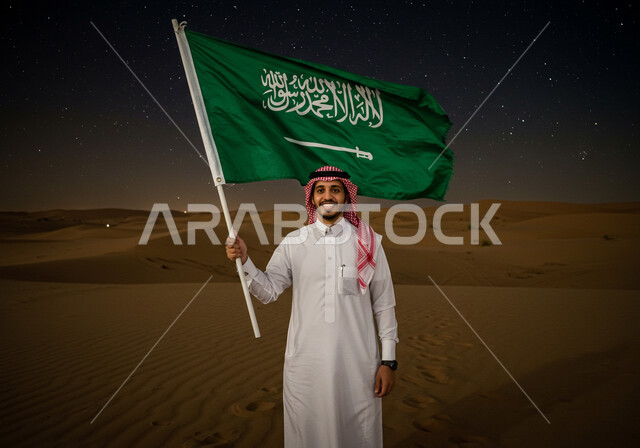 اليوم الوطني السعودي 23 سبتمبر, البيئة الصحراوية في الليل، يوم العلم 11 مارس، رجل عربي خليجي سعودي يرتدي الثوب التقليدي والشماغ يقف على الرمال الناعمة في الصحراء يحمل علم المملكة ينظر إلى الكاميرا بإيماءات الفخر، التشكلات الرملية في الصحاري، خلفية السماء المليئة بالنجوم