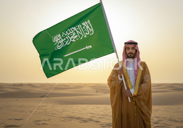 ارتداء الزي الشعبي في مناسبة وطنية ، رجل عربي خليجي سعودي يرتدي البشت والشماغ يقف على الرمال الذهبية في الصحراء ويمسك علم المملكة العربية السعودية، اليوم الوطني السعودي 23 سبتمبر, يوم العلم 11 مارس، ذكرى تاسيس الدوله السعوديه الاولى ١٧٢٧ م،  يوم بدينا ٢٢ فبراير