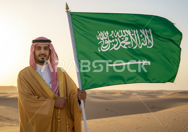 ارتداء الزي الشعبي في مناسبة وطنية ، رجل عربي خليجي سعودي يرتدي البشت والشماغ يقف على الرمال الذهبية في الصحراء ويمسك علم المملكة العربية السعودية، اليوم الوطني السعودي 23 سبتمبر, يوم العلم 11 مارس، ذكرى تاسيس الدوله السعوديه الاولى ١٧٢٧ م،  يوم بدينا ٢٢ فبراير
