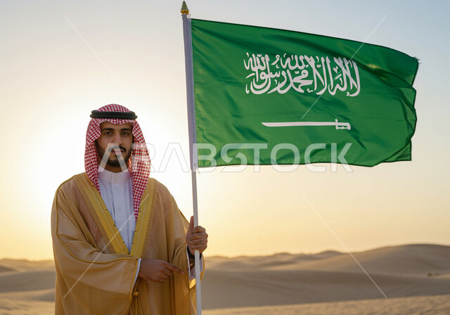 ذكرى تاسيس الدوله السعوديه الاولى ١٧٢٧ م،  يوم بدينا ٢٢ فبراير،  ارتداء الزي الشعبي في مناسبة وطنية، رجل عربي خليجي سعودي يرتدي البشت والشماغ يقف على الرمال الذهبية في الصحراء ويمسك علم المملكة العربية السعودية، اليوم الوطني السعودي 23 سبتمبر, يوم العلم 11 مارس