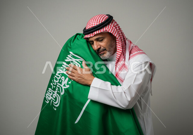 الاحتفال بالمناسبات الوطنية, بورتريه مقرب لشايب عربي خليجي سعودي يرتدي الثوب والشماغ  يحضن علم المملكة العربية السعودية, ايماءات الحب والاعتزاز بالوطن, يوم بدينا ٢٢ فبراير، ذكرى تاسيس الدوله السعوديه الاولى ١٧٢٧ م، خلفية رمادي