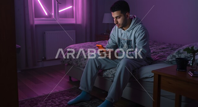 مفهوم الانزعاج لتلقي خبر سيء، استخدام التكنولوجيا والتقنية الحديثة المتطورة، شاب عربي خليجي سعودي يجلس على سرير مريح يمسك بيده الهاتف المحمول يقوم بتصفح تطبيقات التواصل الاجتماعي بإيماءات الملل والقلق، الدردشة مع الأهل والأصدقاء
