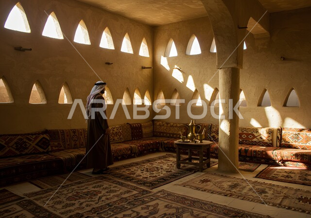 مفهوم الأناقة والاهتمام بالمظهر الخارجي، البيوت التراثية على الطراز التاريخي العريق، رجل عربي خليجي سعودي يرتدي الثوب التقليدي والشماغ يقف باستقامة وسط غرفة طينية قديمة ينظر إلى شيء ما، دخول أشعة الشمس إلى البيت