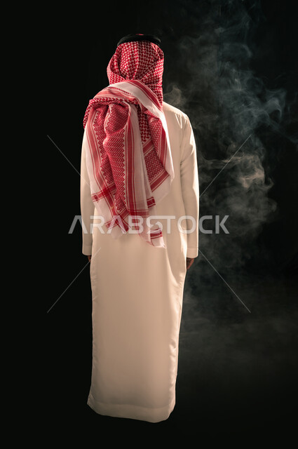 Concept of masculinity and elegance, concern for outward appearance, portrait from behind of a Saudi Arabian Gulf man wearing traditional thobe and shemagh standing straight looking at something, full length body, black background
