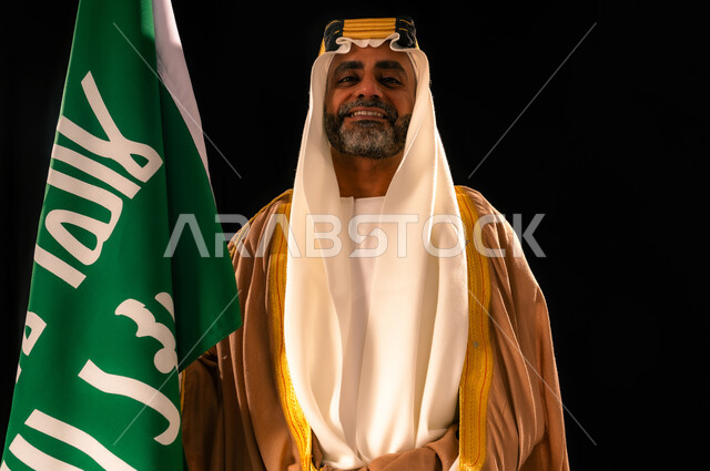 يوم بدينا 22 فبراير، الفخر والاعتزاز بإنجازات المملكة، إظهار الولاء والحب للوطن, بورتريه مقرب لرجل عربي خليجي سعودي يرتدي الغترة والعقال الذهبي والبشت يحمل علم التأسيس ينظر إلى الكاميرا بإيماءات الشموخ، ذكرى تأسيس الدولة السعودية الأولى 1727م، خلفية سوداء