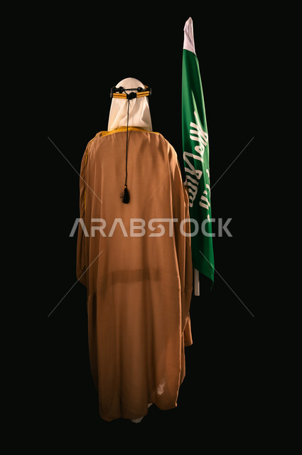 ذكرى تأسيس الدولة السعودية الأولى 1727م، الفخر والاعتزاز بإنجازات المملكة، يوم بدينا 22 فبراير، إظهار الولاء والحب للوطن, بورتريه من الخلف لرجل عربي خليجي سعودي يرتدي الغترة والعقال الذهبي والبشت يحمل علم التأسيس، الجسم بالطول الكامل، خلفية سوداء