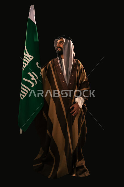 الفخر والاعتزاز بإنجازات المملكة، يوم بدينا 22 فبراير، إظهار الولاء والحب للوطن, ذكرى تأسيس الدولة السعودية الأولى 1727م، بورتريه لرجل عربي خليجي سعودي يرتدي الغترة والبشت يحمل علم التأسيس ينظر إلى شيء ما بإيماءات الشموخ، الجسم بالطول الكامل، خلفية سوداء